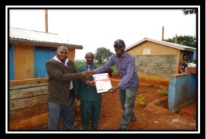 Dan Otieno presents a plaque to Michael Gichira of the Murera group outside the latrines constructed in 2012 with funding from CanAssist.